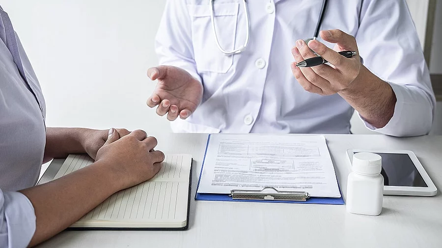 Arzt sitzt seiner Patienten gegenüber und erklärt. Auf dem Tisch vor ihm liegen Dokumente und ein Wasserglas.