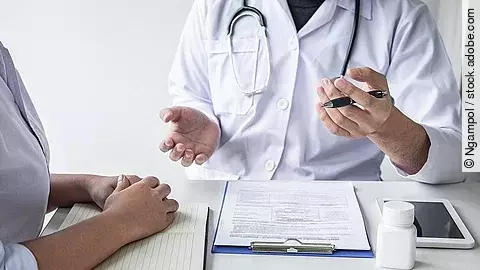 Arzt sitzt seiner Patienten gegenüber und erklärt. Auf dem Tisch vor ihm liegen Dokumente und ein Wasserglas.