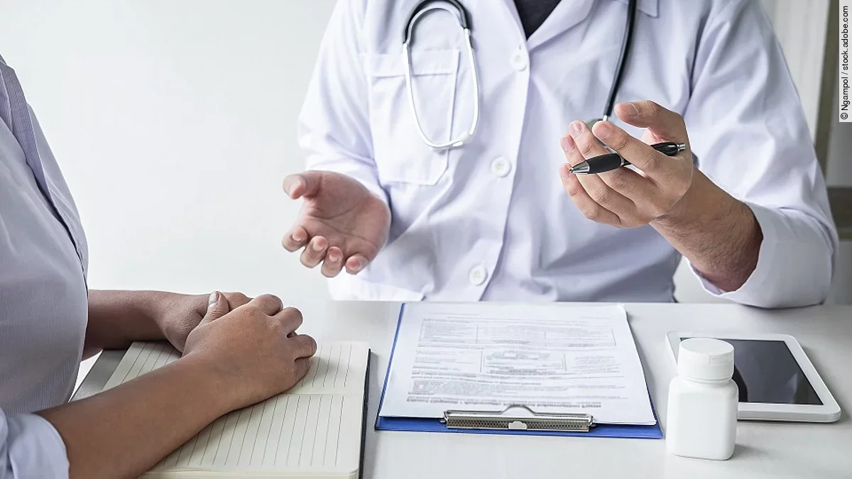 Arzt sitzt seiner Patienten gegenüber und erklärt. Auf dem Tisch vor ihm liegen Dokumente und ein Wasserglas.