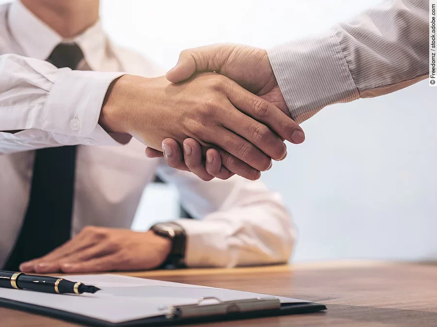 Händeschütteln Ein Mann mit Hemd und Krawatte sitzt an einem Tisch, vor ihm liegt ein Dokument und ein Stift. Er schüttelt die Hand eines anderen Mannes.