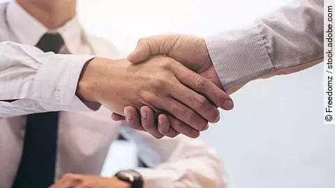 Ein Mann mit Hemd und Krawatte sitzt an einem Tisch, vor ihm liegt ein Dokument und ein Stift. Er schüttelt die Hand eines anderen Mannes.