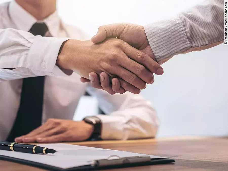 Ein Mann mit Hemd und Krawatte sitzt an einem Tisch, vor ihm liegt ein Dokument und ein Stift. Er schüttelt die Hand eines anderen Mannes.