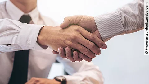 Ein Mann mit Hemd und Krawatte sitzt an einem Tisch, vor ihm liegt ein Dokument und ein Stift. Er schüttelt die Hand eines anderen Mannes.