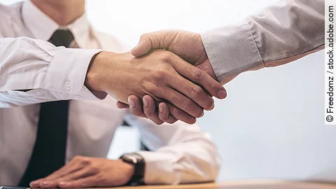 Händeschütteln Ein Mann mit Hemd und Krawatte sitzt an einem Tisch, vor ihm liegt ein Dokument und ein Stift. Er schüttelt die Hand eines anderen Mannes.