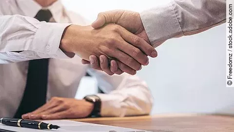 Ein Mann mit Hemd und Krawatte sitzt an einem Tisch, vor ihm liegt ein Dokument und ein Stift. Er schüttelt die Hand eines anderen Mannes.