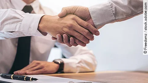 Händeschütteln Ein Mann mit Hemd und Krawatte sitzt an einem Tisch, vor ihm liegt ein Dokument und ein Stift. Er schüttelt die Hand eines anderen Mannes.