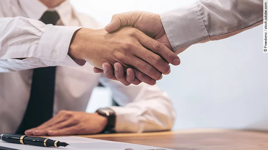 Ein Mann mit Hemd und Krawatte sitzt an einem Tisch, vor ihm liegt ein Dokument und ein Stift. Er schüttelt die Hand eines anderen Mannes.