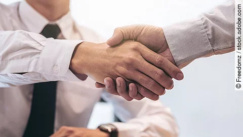 Ein Mann mit Hemd und Krawatte sitzt an einem Tisch, vor ihm liegt ein Dokument und ein Stift. Er schüttelt die Hand eines anderen Mannes.