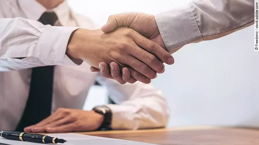 Händeschütteln Ein Mann mit Hemd und Krawatte sitzt an einem Tisch, vor ihm liegt ein Dokument und ein Stift. Er schüttelt die Hand eines anderen Mannes.