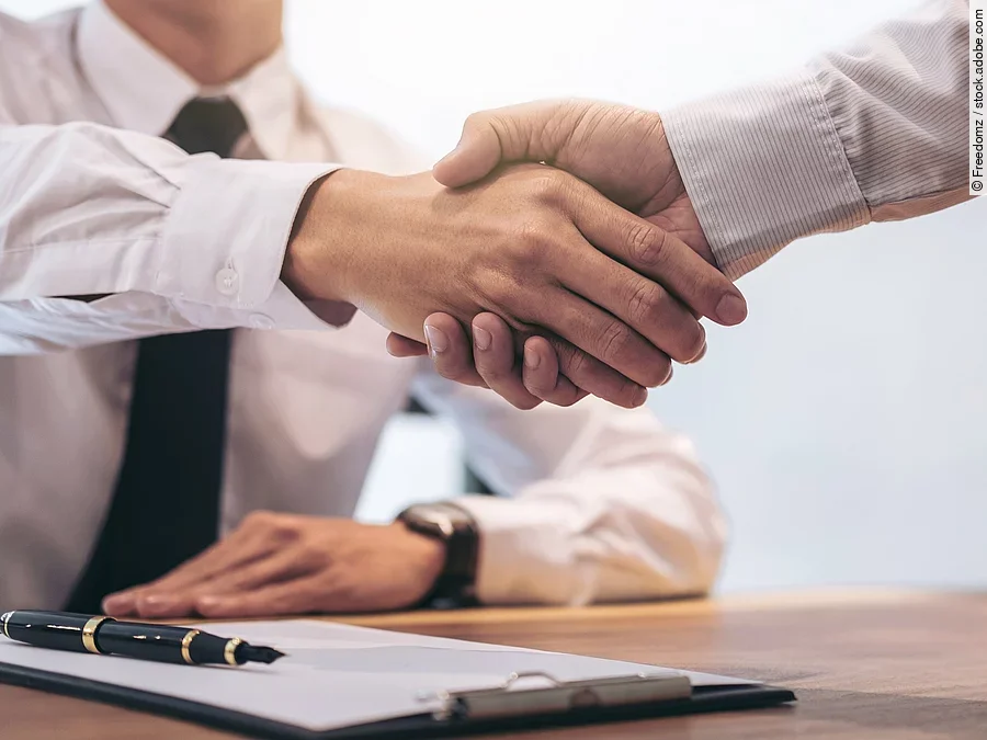 Ein Mann mit Hemd und Krawatte sitzt an einem Tisch, vor ihm liegt ein Dokument und ein Stift. Er schüttelt die Hand eines anderen Mannes.