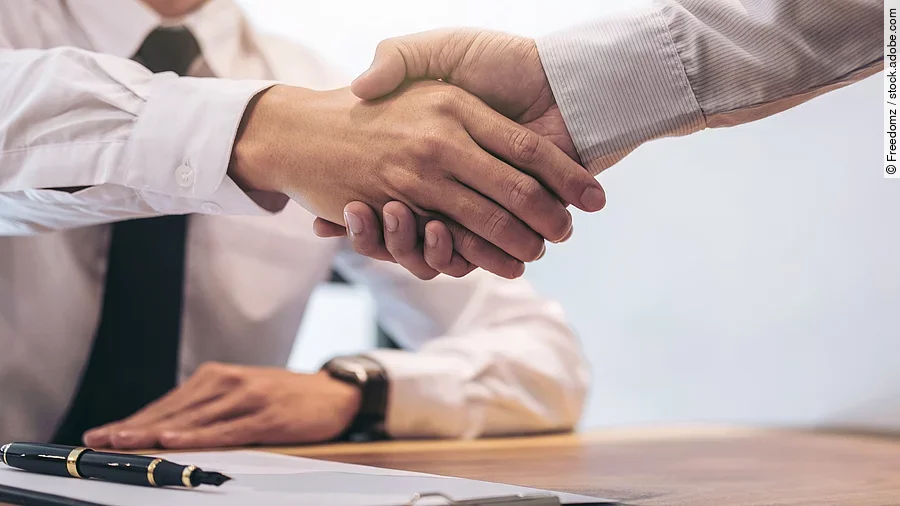 Ein Mann mit Hemd und Krawatte sitzt an einem Tisch, vor ihm liegt ein Dokument und ein Stift. Er schüttelt die Hand eines anderen Mannes.