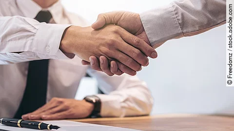 Ein Mann mit Hemd und Krawatte sitzt an einem Tisch, vor ihm liegt ein Dokument und ein Stift. Er schüttelt die Hand eines anderen Mannes.