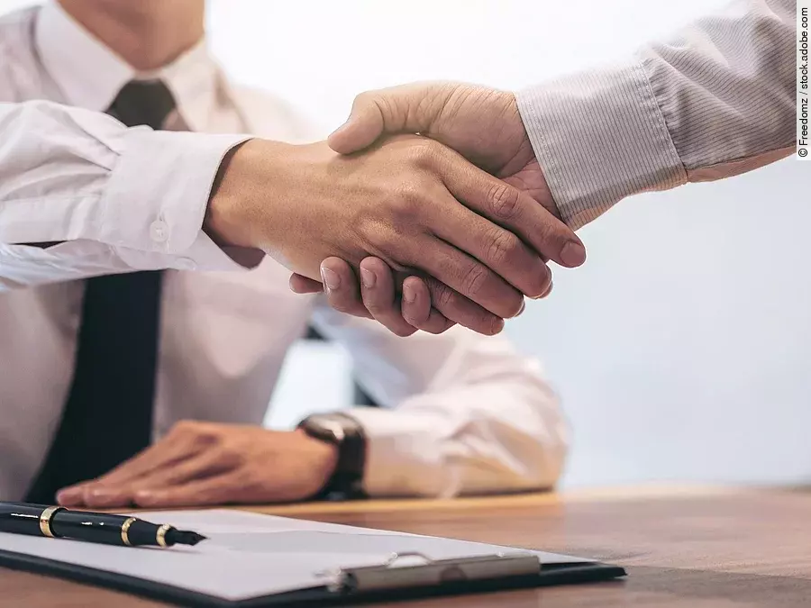 Ein Mann mit Hemd und Krawatte sitzt an einem Tisch, vor ihm liegt ein Dokument und ein Stift. Er schüttelt die Hand eines anderen Mannes.