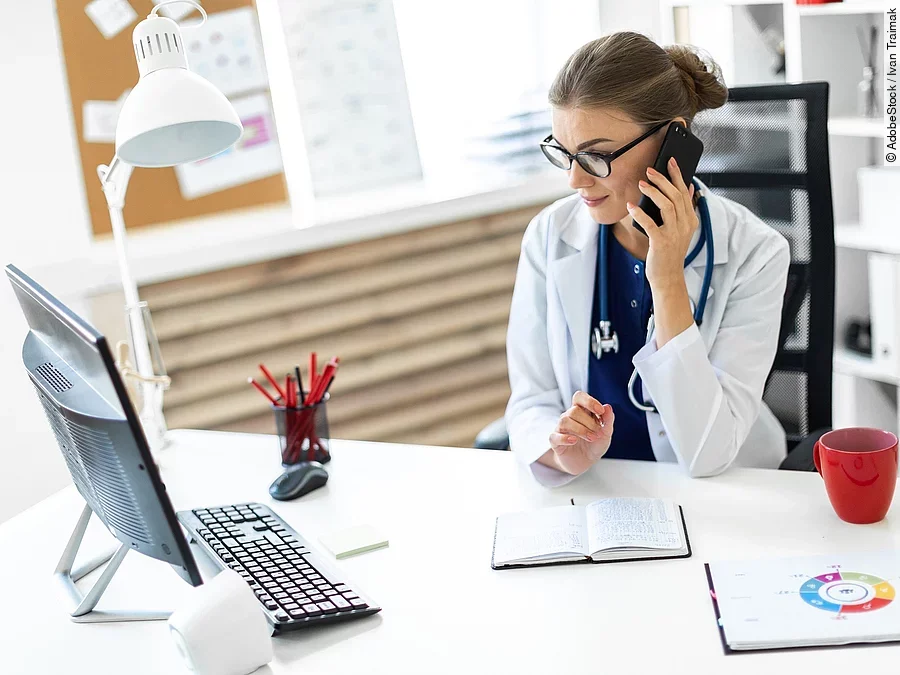 Ärztin sitzt an ihrem Schreibtisch und telefoniert. Vor ihr liegt eine Klemmmappe und ein Bildschirm.