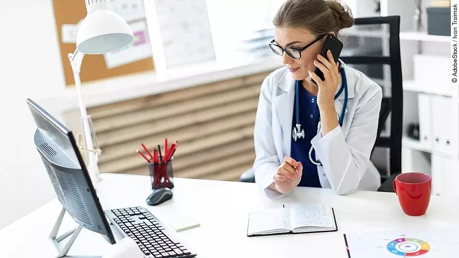 Ärztin sitzt an ihrem Schreibtisch und telefoniert. Vor ihr liegt eine Klemmmappe und ein Bildschirm.