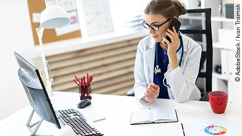 Ärztin sitzt an ihrem Schreibtisch und telefoniert. Vor ihr liegt eine Klemmmappe und ein Bildschirm.