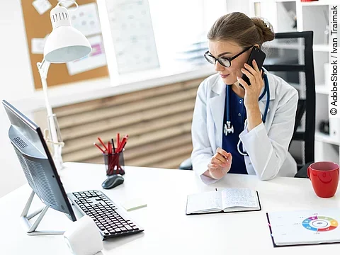 Ärztin sitzt an ihrem Schreibtisch und telefoniert. Vor ihr liegt eine Klemmmappe und ein Bildschirm.