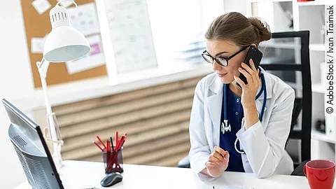 Ärztin sitzt an ihrem Schreibtisch und telefoniert. Vor ihr liegt eine Klemmmappe und ein Bildschirm.
