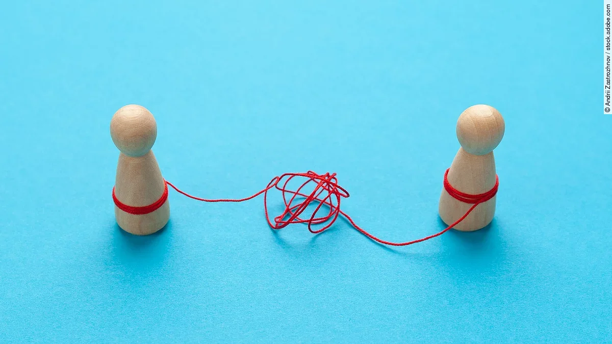 Zwei Holz-Spielfiguren vor blauem Hintergrund stehen sich gegenüber und sind mit einer verknoteten roten Schnur verbunden.