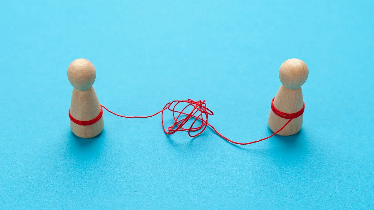 Zwei Holz-Spielfiguren vor blauem Hintergrund stehen sich gegenüber und sind mit einer verknoteten roten Schnur verbunden.