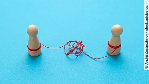 Zwei Holz-Spielfiguren vor blauem Hintergrund stehen sich gegenüber und sind mit einer verknoteten roten Schnur verbunden.