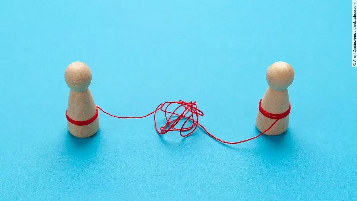 Zwei Holz-Spielfiguren vor blauem Hintergrund stehen sich gegenüber und sind mit einer verknoteten roten Schnur verbunden.
