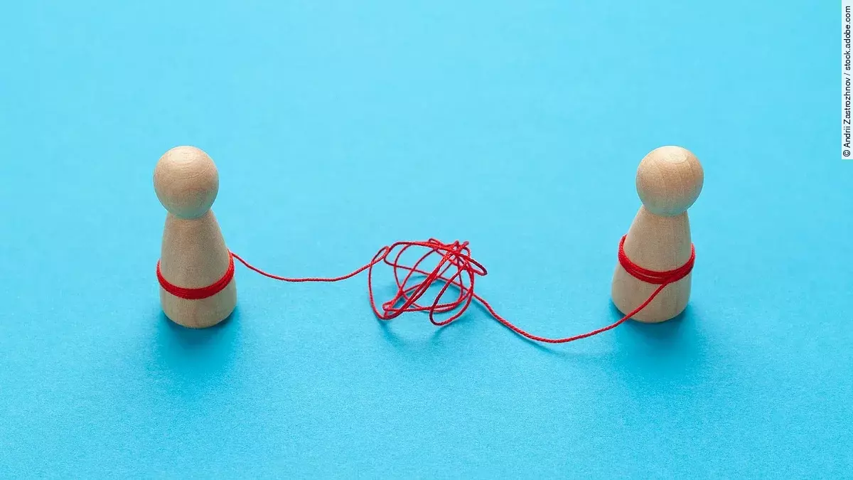 Zwei Holz-Spielfiguren vor blauem Hintergrund stehen sich gegenüber und sind mit einer verknoteten roten Schnur verbunden.