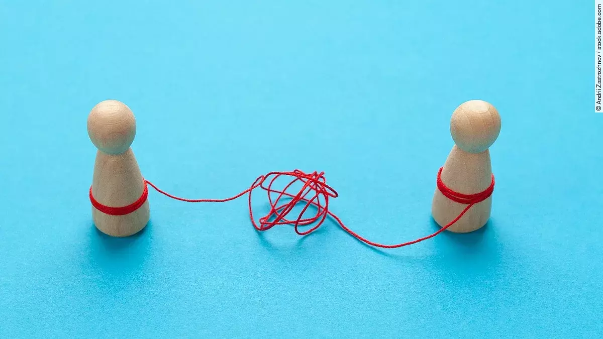 Zwei Holz-Spielfiguren vor blauem Hintergrund stehen sich gegenüber und sind mit einer verknoteten roten Schnur verbunden.