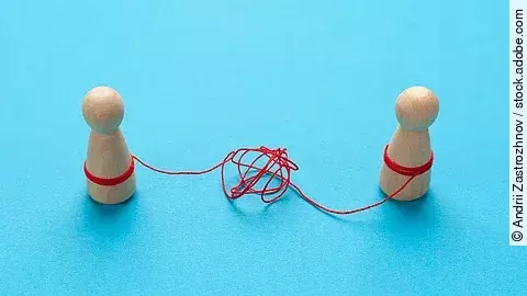 Zwei Holz-Spielfiguren vor blauem Hintergrund stehen sich gegenüber und sind mit einer verknoteten roten Schnur verbunden.