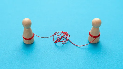 Zwei Holz-Spielfiguren vor blauem Hintergrund stehen sich gegenüber und sind mit einer verknoteten roten Schnur verbunden.