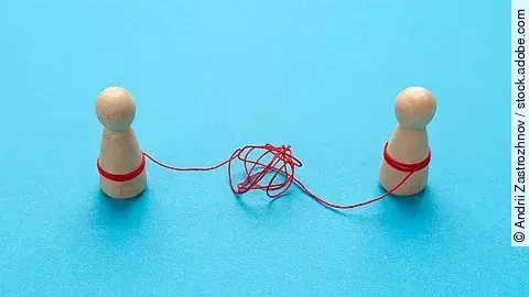 Zwei Holz-Spielfiguren vor blauem Hintergrund stehen sich gegenüber und sind mit einer verknoteten roten Schnur verbunden.