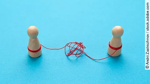 Zwei Holz-Spielfiguren vor blauem Hintergrund stehen sich gegenüber und sind mit einer verknoteten roten Schnur verbunden.
