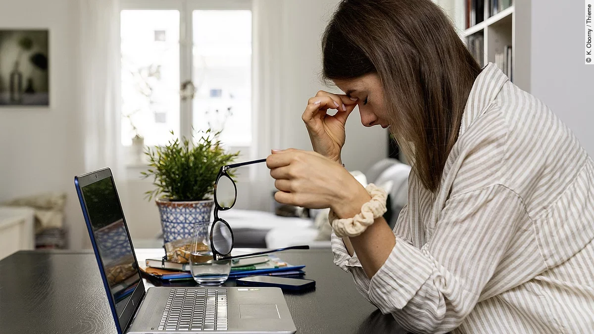 Junge Frau am Laptop sitzend, fasst sich an Nasenwurzel und stützt den Kopf mit der rechten Hand, die linke Hand hält die abgenommene Brille.