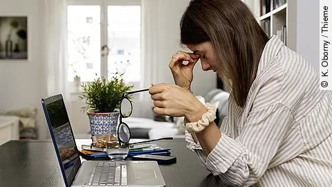 Arbeiten mit Long Covid Junge Frau am Laptop sitzend, fasst sich an Nasenwurzel und stützt den Kopf mit der rechten Hand, die linke Hand hält die abgenommene Brille.
