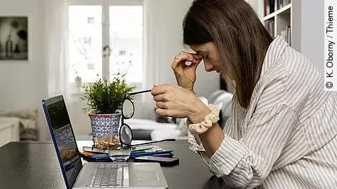 Junge Frau am Laptop sitzend, fasst sich an Nasenwurzel und stützt den Kopf mit der rechten Hand, die linke Hand hält die abgenommene Brille.