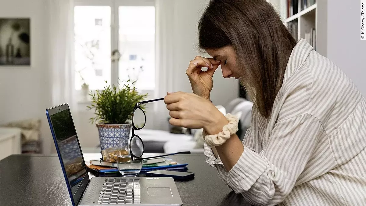 Junge Frau am Laptop sitzend, fasst sich an Nasenwurzel und stützt den Kopf mit der rechten Hand, die linke Hand hält die abgenommene Brille.