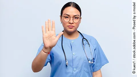 Stop Eine Frau im Kasack und mit Stethoskop schaut ernst und hebt die Hand als Stopzeichen