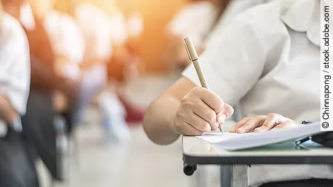 Ein Student schreibt etwas in einen Ordner auf. Hinter ihm sind verschwommen mehrere Reihen mit sitzenden Studierenden zu sehen.