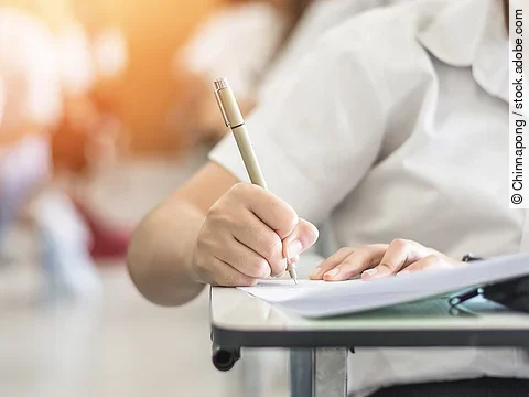 Ein Student schreibt etwas in einen Ordner auf. Hinter ihm sind verschwommen mehrere Reihen mit sitzenden Studierenden zu sehen.