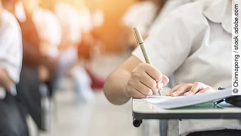 Studium Ein Student schreibt etwas in einen Ordner auf. Hinter ihm sind verschwommen mehrere Reihen mit sitzenden Studierenden zu sehen.