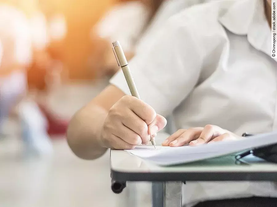 Ein Student schreibt etwas in einen Ordner auf. Hinter ihm sind verschwommen mehrere Reihen mit sitzenden Studierenden zu sehen.