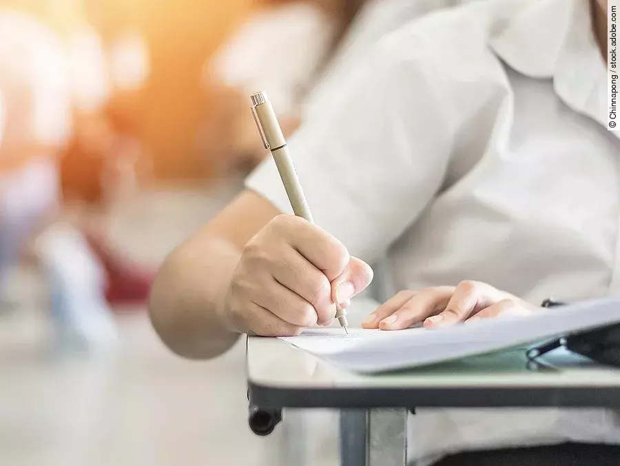 Studium Ein Student schreibt etwas in einen Ordner auf. Hinter ihm sind verschwommen mehrere Reihen mit sitzenden Studierenden zu sehen.