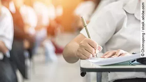 Ein Student schreibt etwas in einen Ordner auf. Hinter ihm sind verschwommen mehrere Reihen mit sitzenden Studierenden zu sehen.