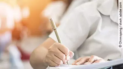 Ein Student schreibt etwas in einen Ordner auf. Hinter ihm sind verschwommen mehrere Reihen mit sitzenden Studierenden zu sehen.