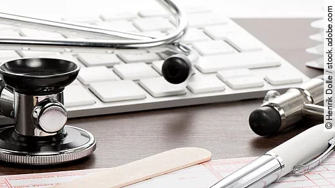 Rezept Auf einem Tisch liegt ein rosafarbenes Rezept, darauf und daneben verteilt liegen ein Stethoskop, ein Kugelschreiber, ein Holzspatel und eine Tastatur.