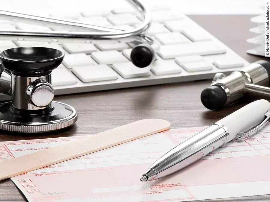 Auf einem Tisch liegt ein rosafarbenes Rezept, darauf und daneben verteilt liegen ein Stethoskop, ein Kugelschreiber, ein Holzspatel und eine Tastatur.