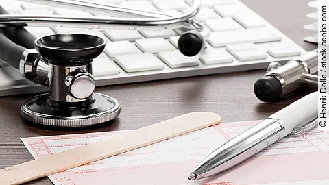 Auf einem Tisch liegt ein rosafarbenes Rezept, darauf und daneben verteilt liegen ein Stethoskop, ein Kugelschreiber, ein Holzspatel und eine Tastatur.