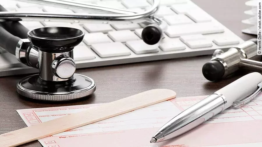 Auf einem Tisch liegt ein rosafarbenes Rezept, darauf und daneben verteilt liegen ein Stethoskop, ein Kugelschreiber, ein Holzspatel und eine Tastatur.