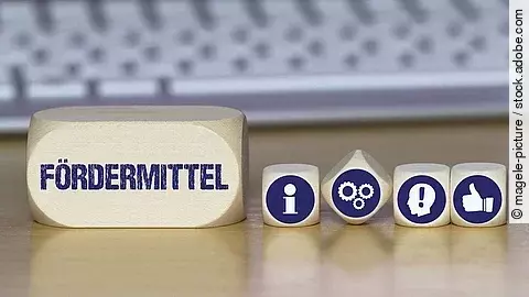 Ein Holzklotz mit der Aufschrift Fördermittel. Daneben liegen Würfel mit den Symbolen i (für Information), Zahnrädchen, ein Kopf mit einem Ausrufezeichen und ein Daumen-hoch.