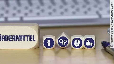 Ein Holzklotz mit der Aufschrift Fördermittel. Daneben liegen Würfel mit den Symbolen i (für Information), Zahnrädchen, ein Kopf mit einem Ausrufezeichen und ein Daumen-hoch.